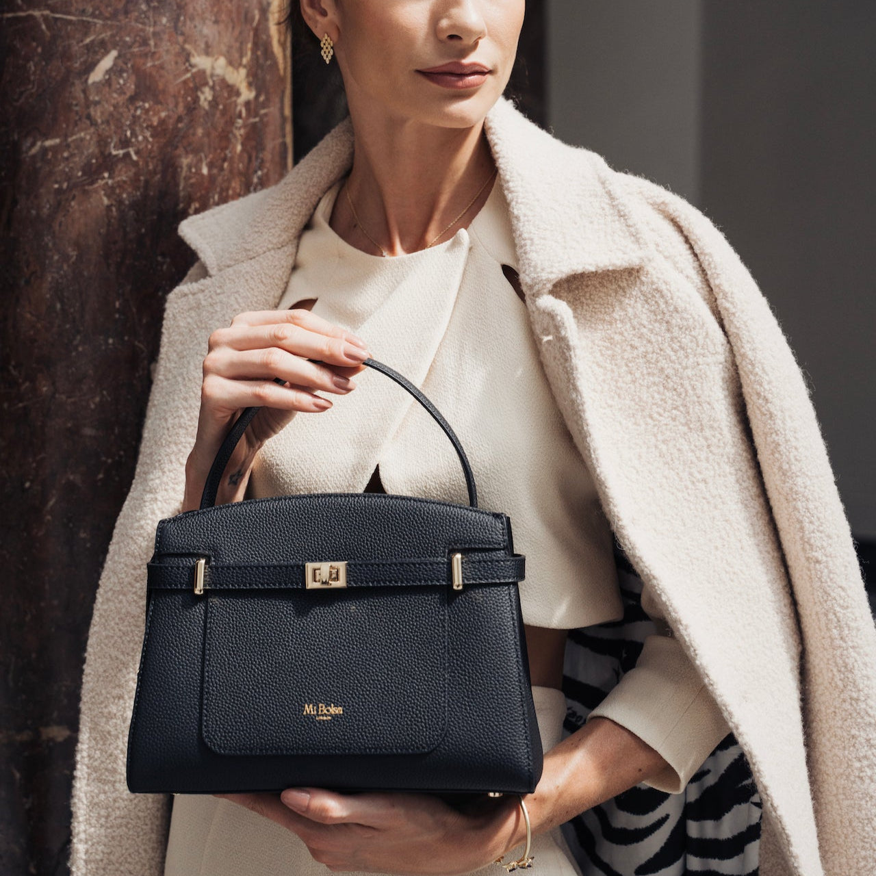 Woman holding a black Mi Bolsa Manhattan Bag against a textured wall