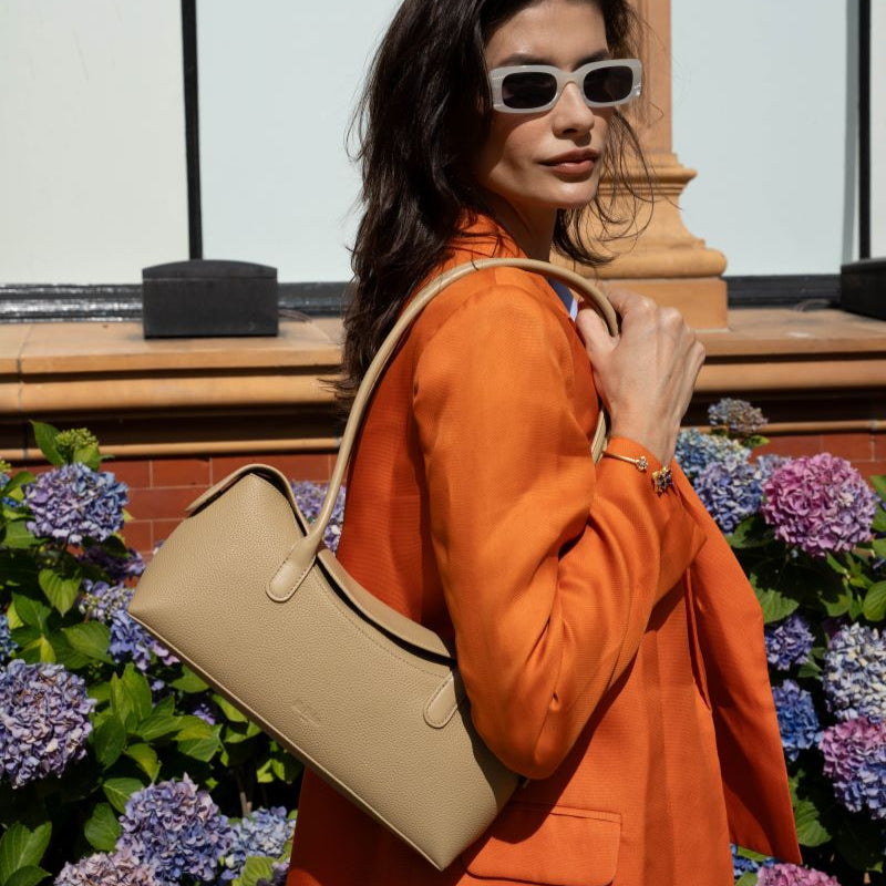 Woman in an orange coat holding a Mi Bolsa Rosa handbag with flowers in the background