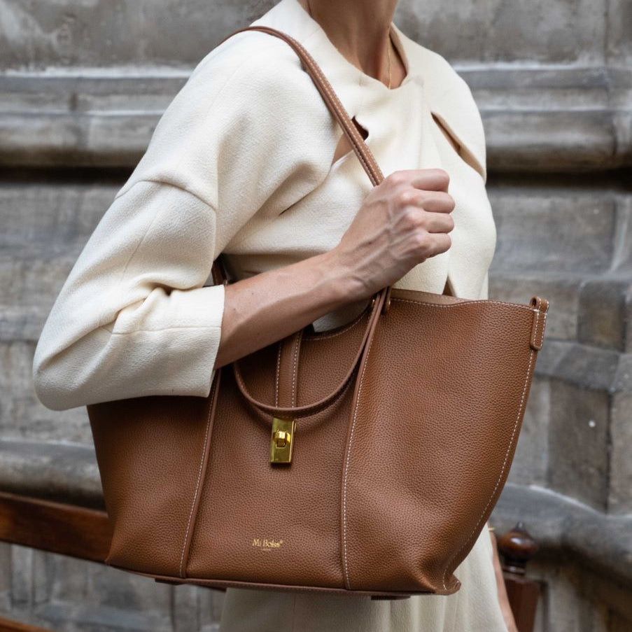 A woman holding a brown Mi Bolsa Wharf handbag against a textured wall background