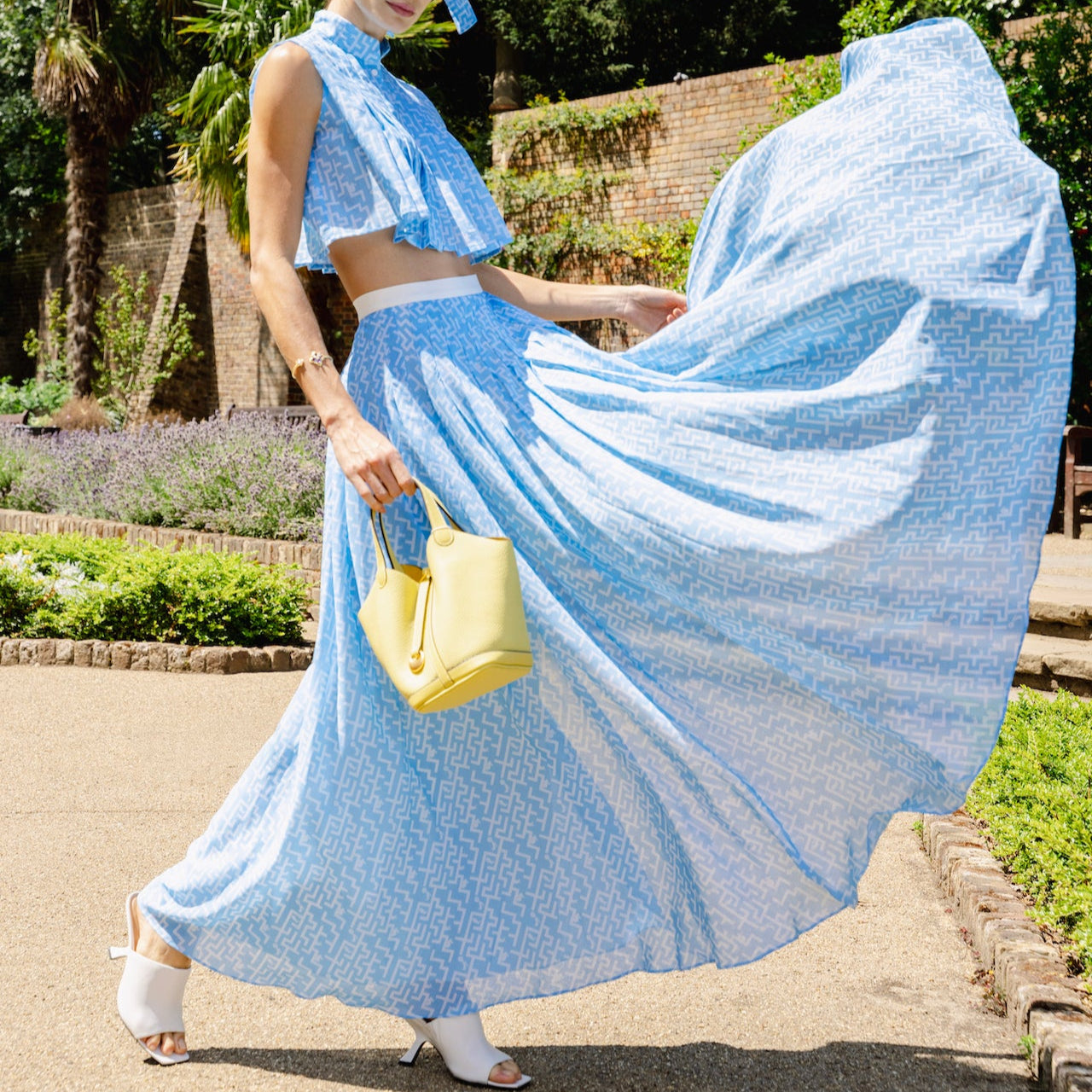 A woman in a blue dress with a Mi Bolsa Monaco Basket handbag standing outdoors in a garden setting.
